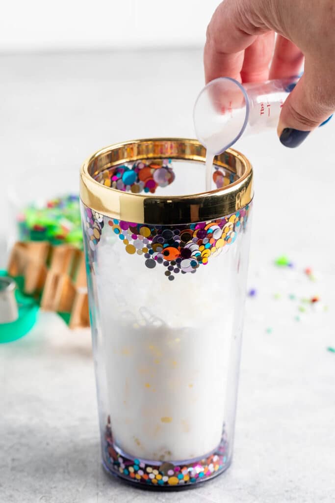 A clear tumbler with colorful circular decorations and a gold rim is filled with a creamy white liquid. A hand pours more of the liquid from a small measuring cup into the tumbler. Some confetti is scattered on the gray surface.