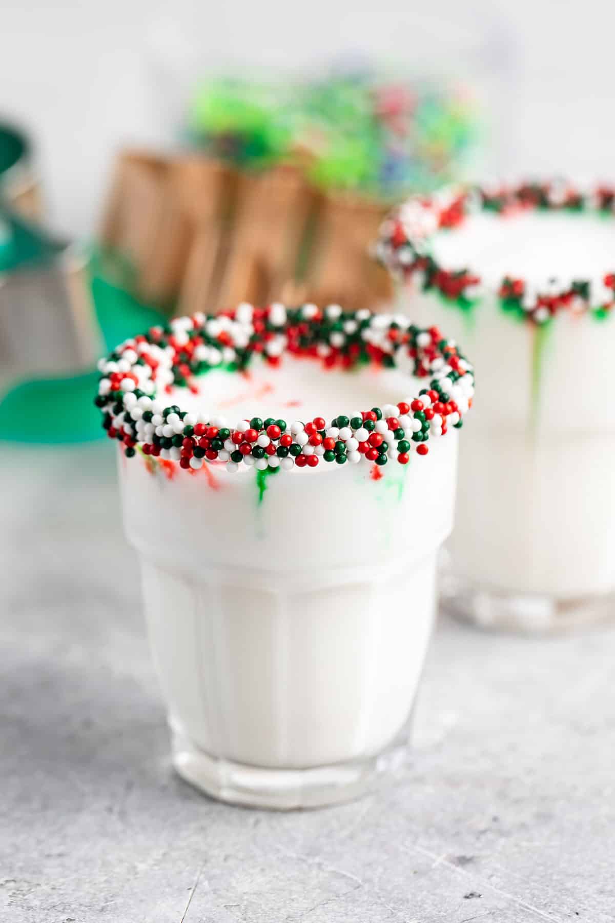 A glass of milk with a festive rim coated in red, green, and white sprinkles. Another similar glass is partially visible in the background. The setting suggests a holiday theme with blurred decorations in soft focus.