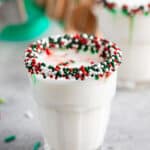 A glass of milk with a rim coated in red, green, and white sprinkles sits on a light-colored surface. A second glass and blurred objects are in the background, suggesting a festive atmosphere.