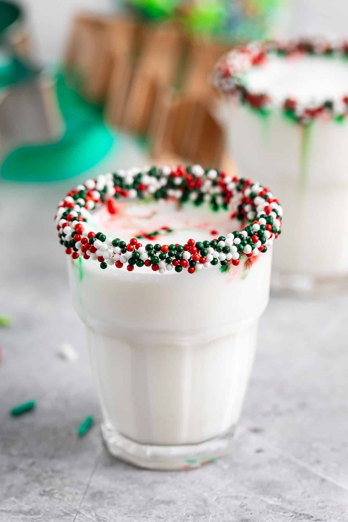 A glass of milk with a rim coated in red, green, and white sprinkles sits on a light-colored surface. A second glass and blurred objects are in the background, suggesting a festive atmosphere.