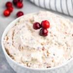 A white bowl filled with creamy cranberry salad, topped with three whole cranberries. Nearby, more cranberries are scattered on a light gray surface, and a striped cloth is partially visible in the background.