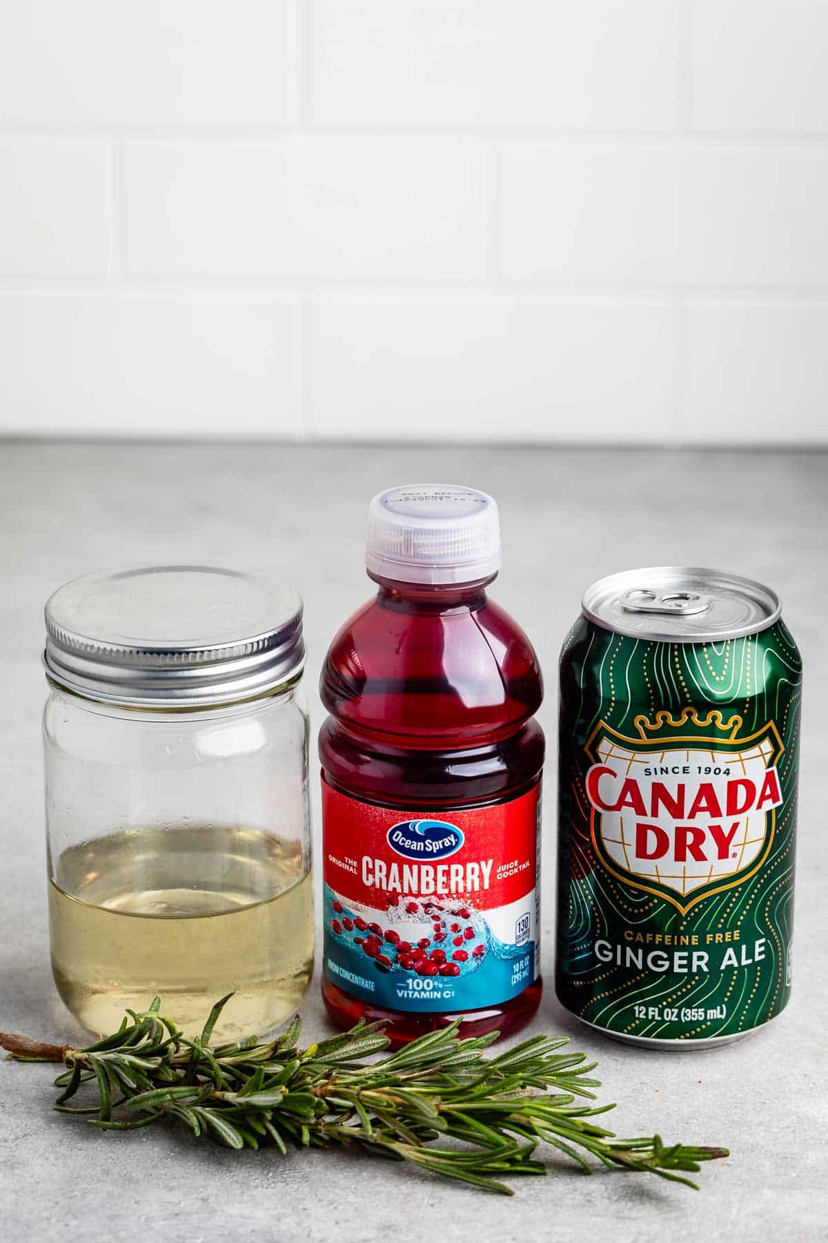 A mason jar filled with a clear liquid, a bottle of cranberry juice, a can of Canada Dry ginger ale, and a sprig of rosemary are arranged on a countertop with a white tiled background.