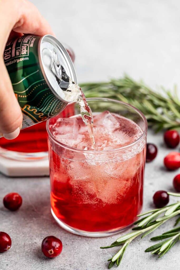 A hand pouring sparkling beverage from a can into a glass filled with ice and red liquid. Fresh cranberries and rosemary sprigs are scattered around on a gray surface.