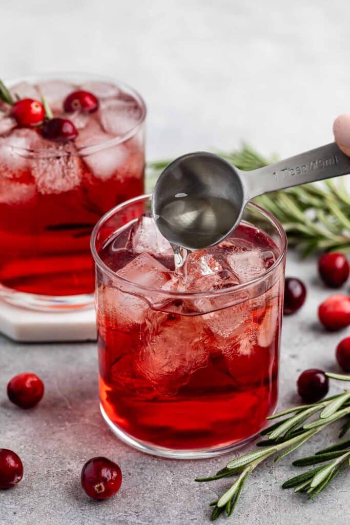 Two glasses filled with red cranberry cocktails, garnished with rosemary and cranberries. A tablespoon measures liquid into one glass. Fresh cranberries and rosemary sprigs are scattered on a light gray surface.