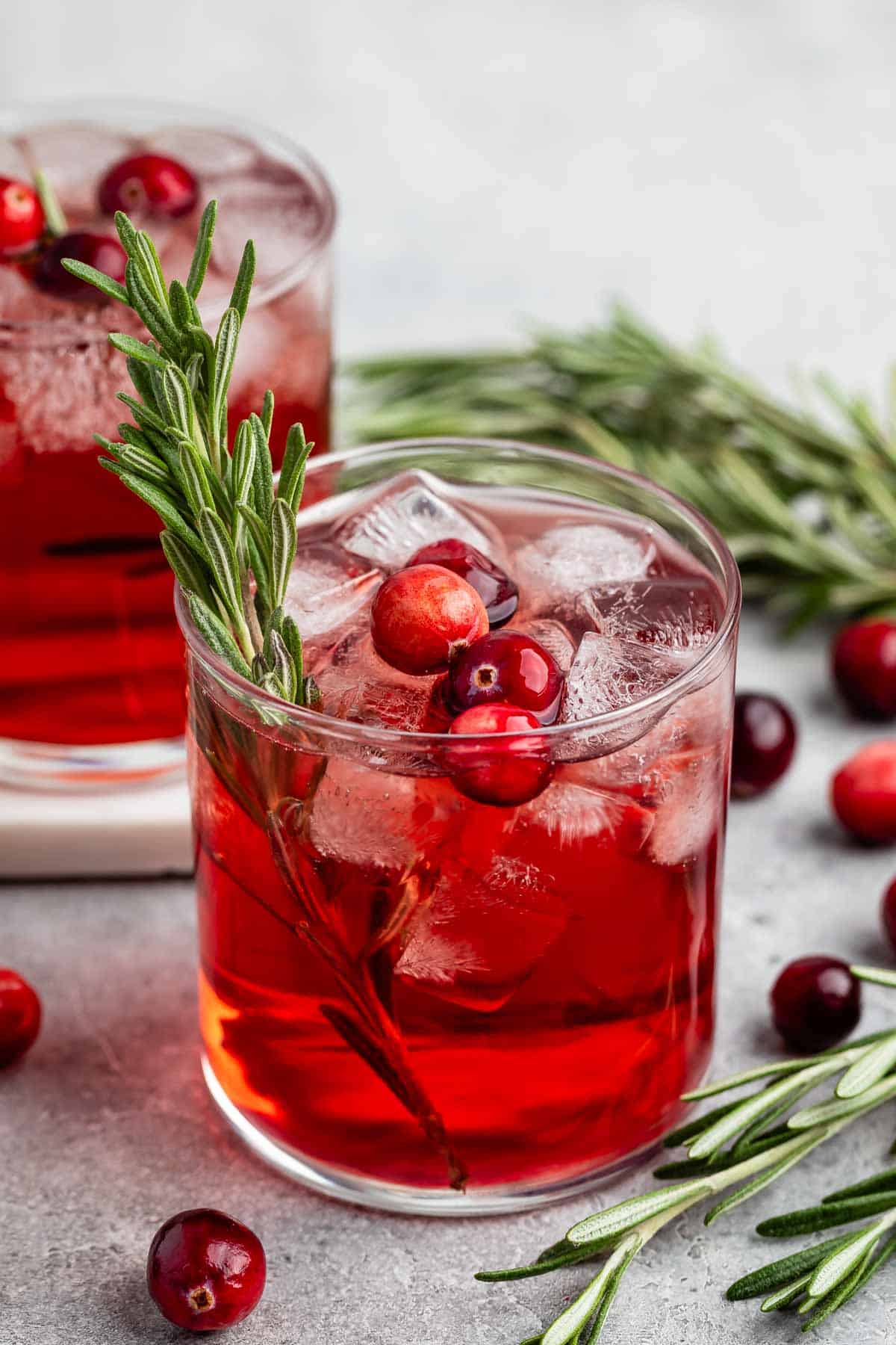 A glass of red cranberry cocktail with ice cubes, garnished with fresh cranberries and a sprig of rosemary. Another similar drink is slightly blurred in the background, with loose cranberries and rosemary scattered on a gray surface.