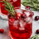 A refreshing cranberry cocktail in a glass filled with ice cubes, garnished with fresh rosemary and cranberries. Another glass and sprigs of rosemary are blurred in the background.