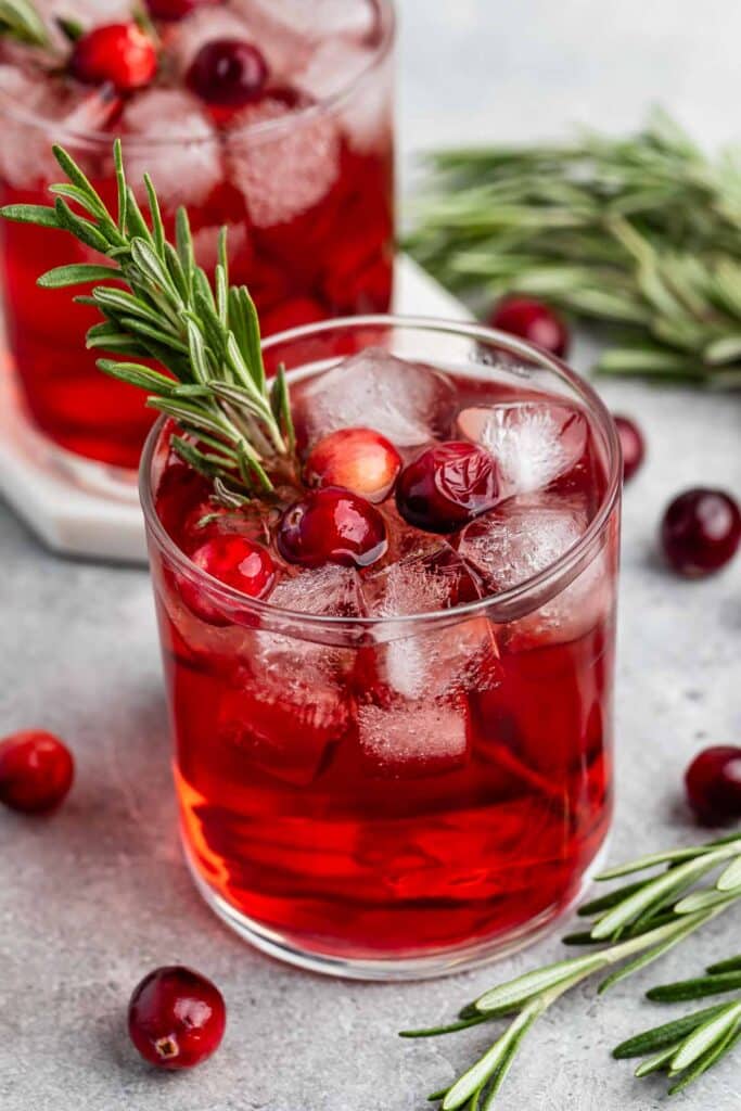 A refreshing cranberry cocktail in a glass filled with ice cubes, garnished with fresh rosemary and cranberries. Another glass and sprigs of rosemary are blurred in the background.