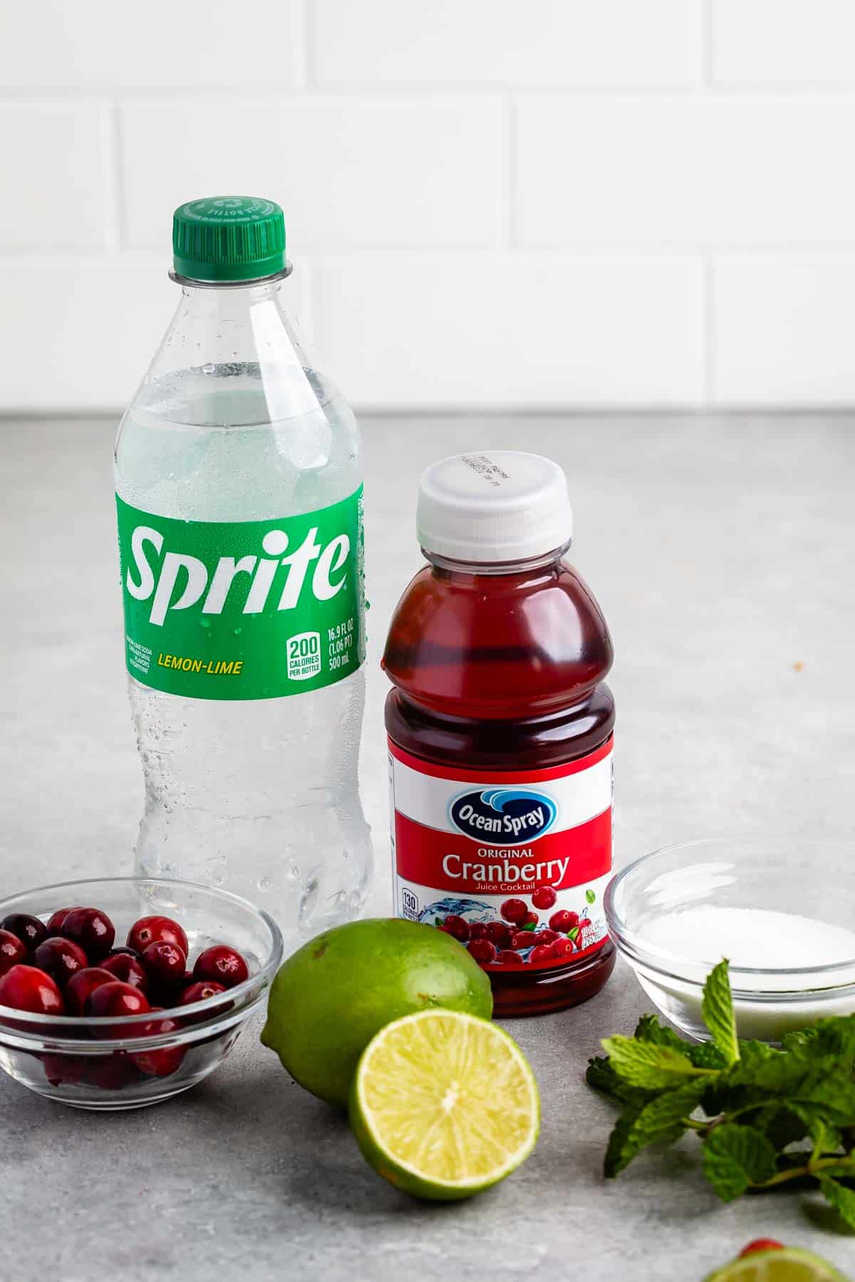 A bottle of Sprite and a bottle of Ocean Spray cranberry juice are on a counter, surrounded by fresh cranberries, a halved lime, a small bowl of sugar, and sprigs of mint. A white tiled wall serves as the background.