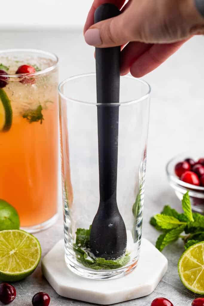 A hand using a muddler in an empty glass with mint leaves at the bottom. Next to the glass are fresh lime halves, mint, and cranberries. In the background, theres a completed drink garnished with cranberries and mint.