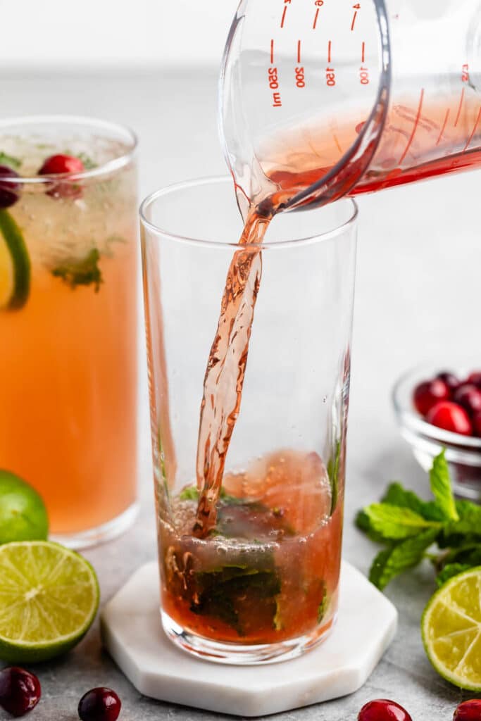 A red liquid is being poured from a measuring cup into a glass containing mint leaves. A lime slice, cranberries, and mint leaves are nearby. Another beverage with cranberries and a sprig of mint is in the background.