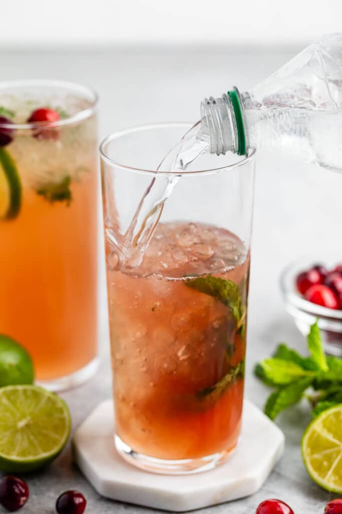 A close-up of a refreshing beverage being poured into a tall glass with ice, mint leaves, and a reddish liquid. Nearby are cranberries, sliced lime, and another similar drink garnished with mint and cranberries.