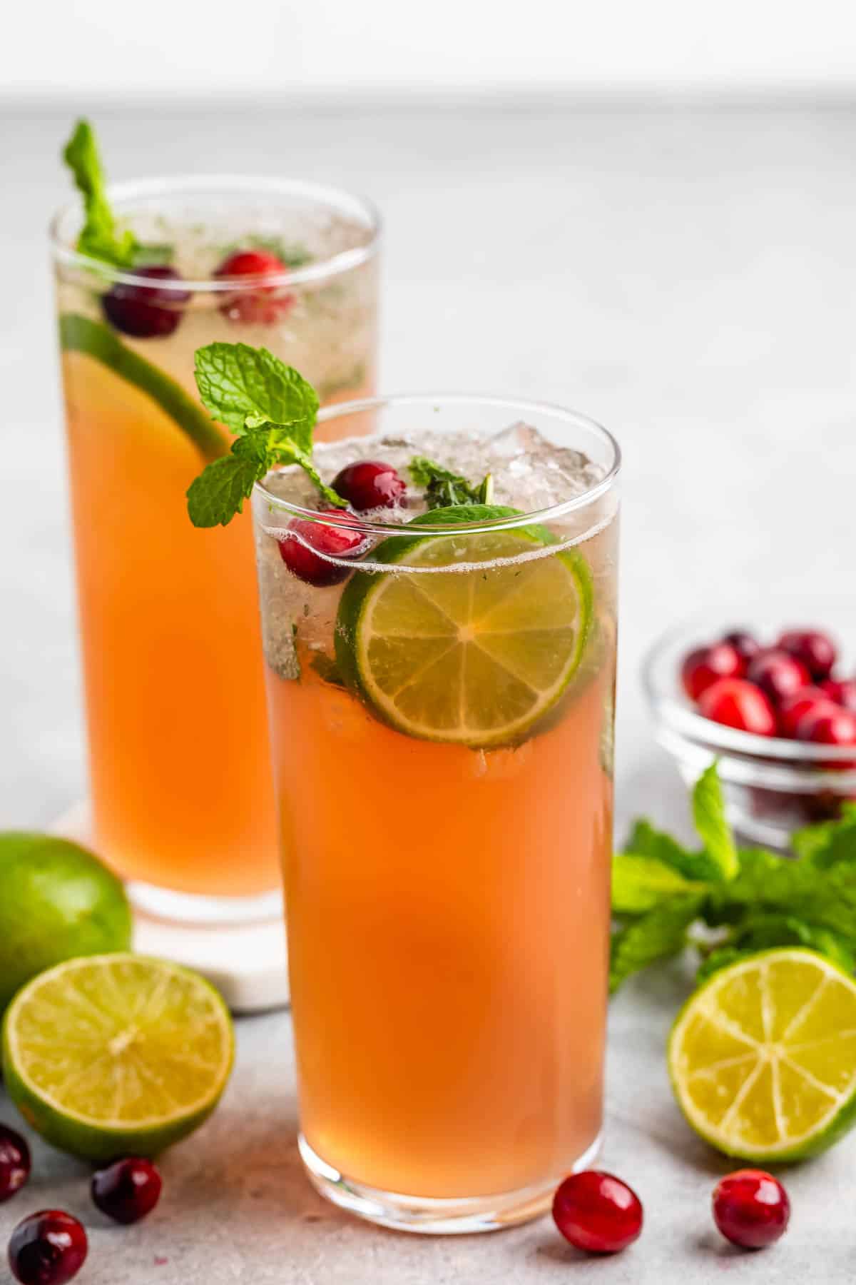 Two tall glasses filled with a pinkish-orange drink, garnished with lime slices, mint leaves, and cranberries. A bowl of cranberries, lime halves, and mint sprigs are nearby on a light surface.