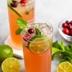 Two tall glasses of refreshing cranberry mojito garnished with lime slices, mint leaves, and cranberries. A few limes and cranberries are scattered on the table nearby. Clear ice cubes float at the top of the drinks.