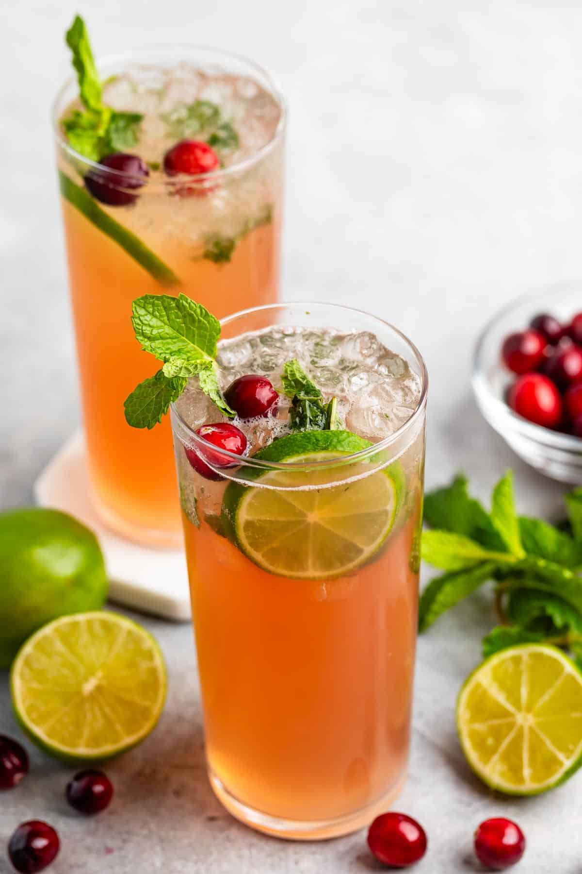 Two tall glasses of refreshing cranberry mojito garnished with lime slices, mint leaves, and cranberries. A few limes and cranberries are scattered on the table nearby. Clear ice cubes float at the top of the drinks.