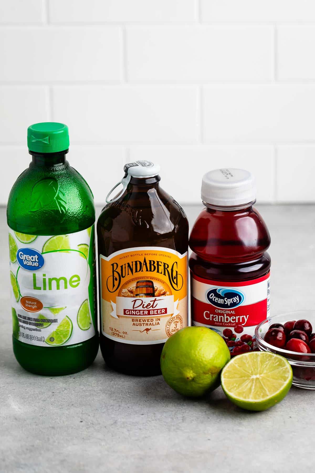 Bottles of lime juice, diet ginger beer, and cranberry juice are displayed on a countertop. Fresh cranberries and a sliced lime sit in front of the bottles, with a white tiled wall in the background.