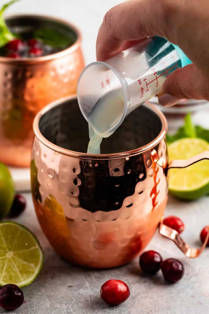 A hand pours liquid from a small measuring cup into a copper mug. Lime and cranberries are scattered around. Another copper mug with cranberries and mint in the background.