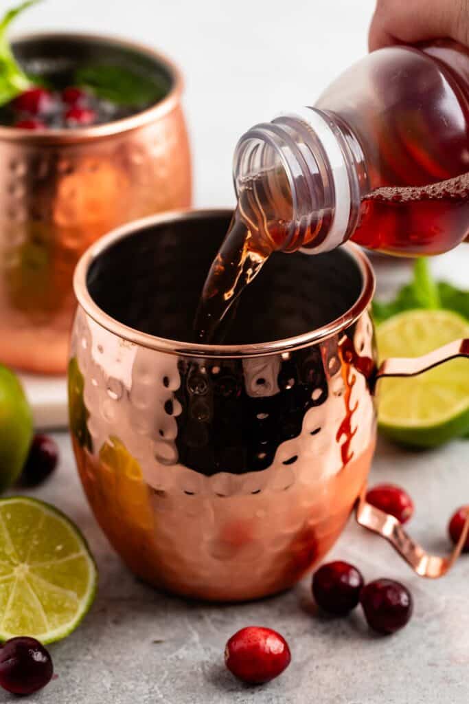 A copper mug is being filled with a red liquid from a bottle. Another copper mug, cranberries, lime halves, and mint leaves surround the scene, suggesting a festive cocktail preparation.