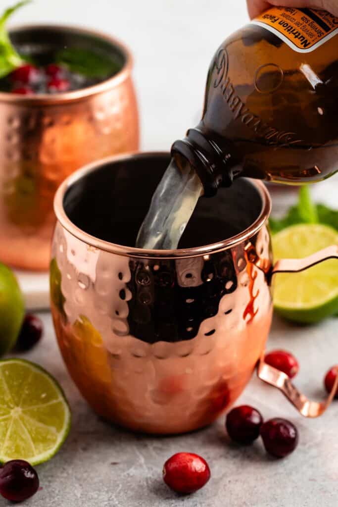 Pouring ginger beer into a copper mug surrounded by lime halves, cranberries, and mint leaves. A second mug is partially visible in the background.