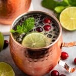 A copper mug filled with a dark drink, garnished with fresh mint, a lime slice, and cranberries. Additional cranberries and lime slices are scattered around the mug on a light surface. Another mug and a bowl of cranberries are visible in the background.