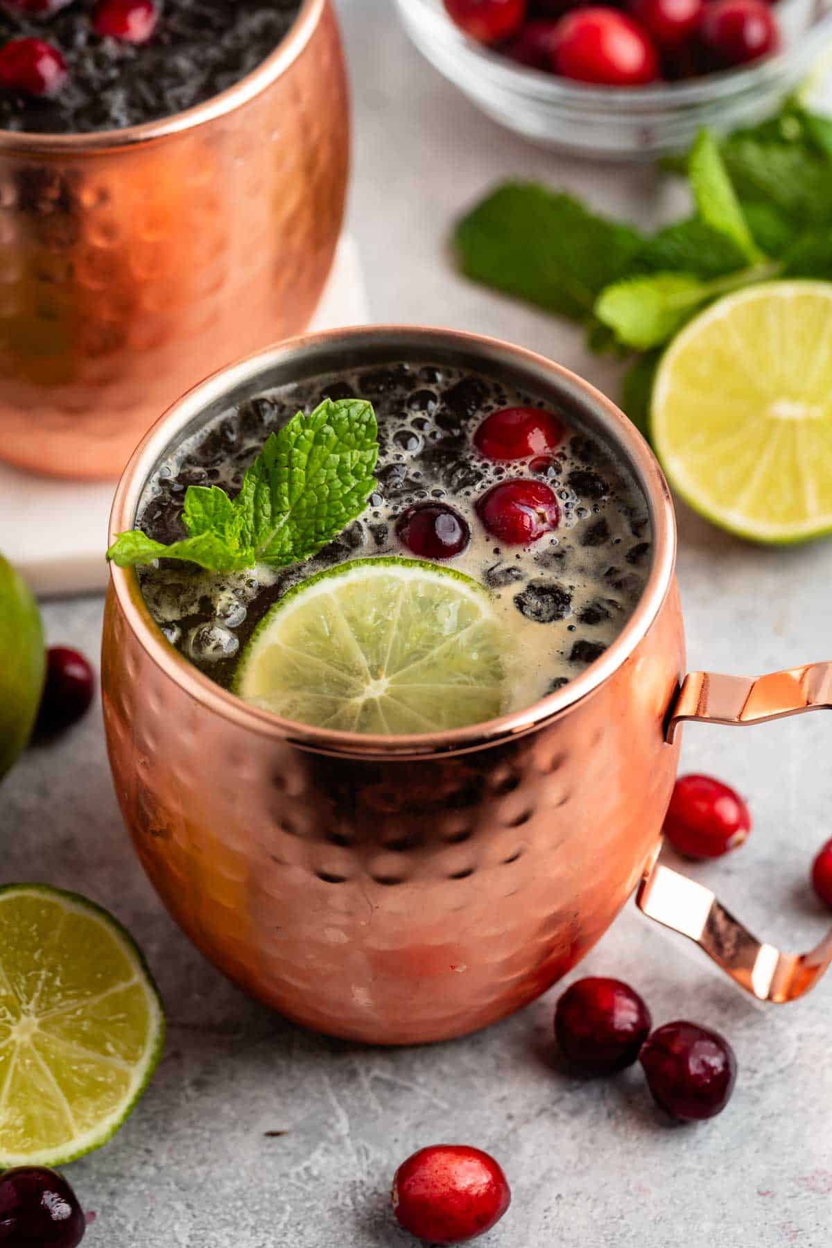 A copper mug filled with a dark drink, garnished with fresh mint, a lime slice, and cranberries. Additional cranberries and lime slices are scattered around the mug on a light surface. Another mug and a bowl of cranberries are visible in the background.