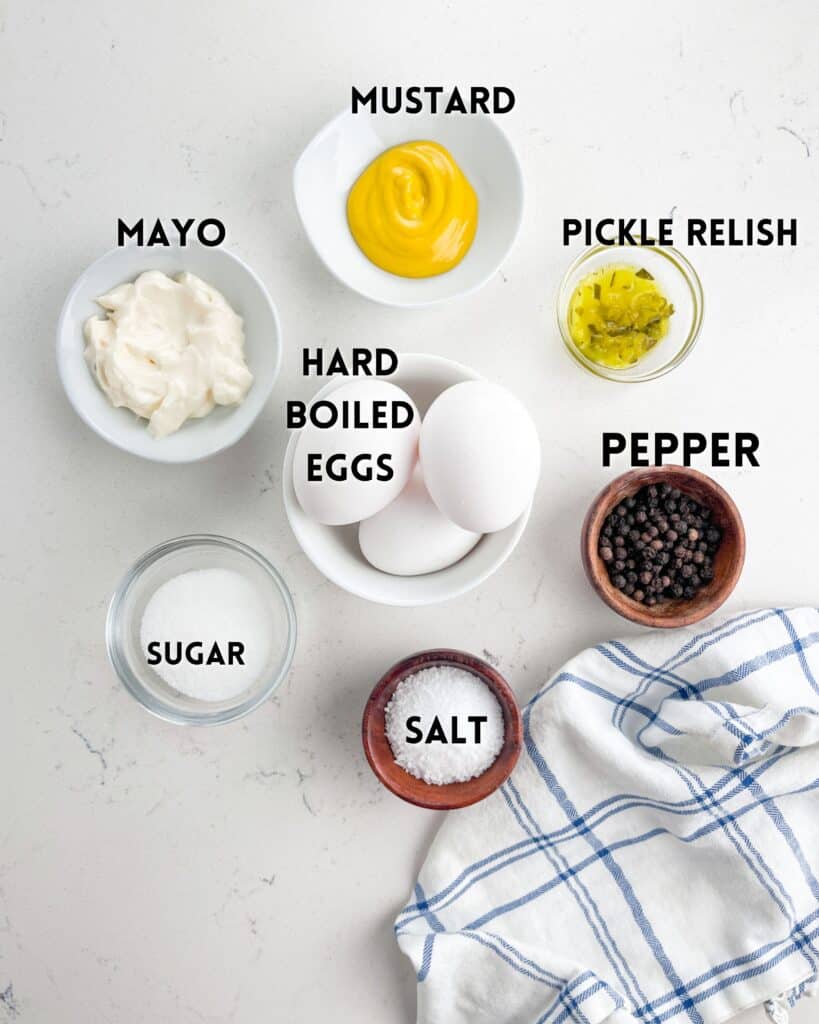 Ingredients for a dish are arranged on a white surface. Includes bowls of mustard, mayo, pickle relish, three hard boiled eggs, sugar, salt, and pepper. A blue and white checkered cloth is nearby.