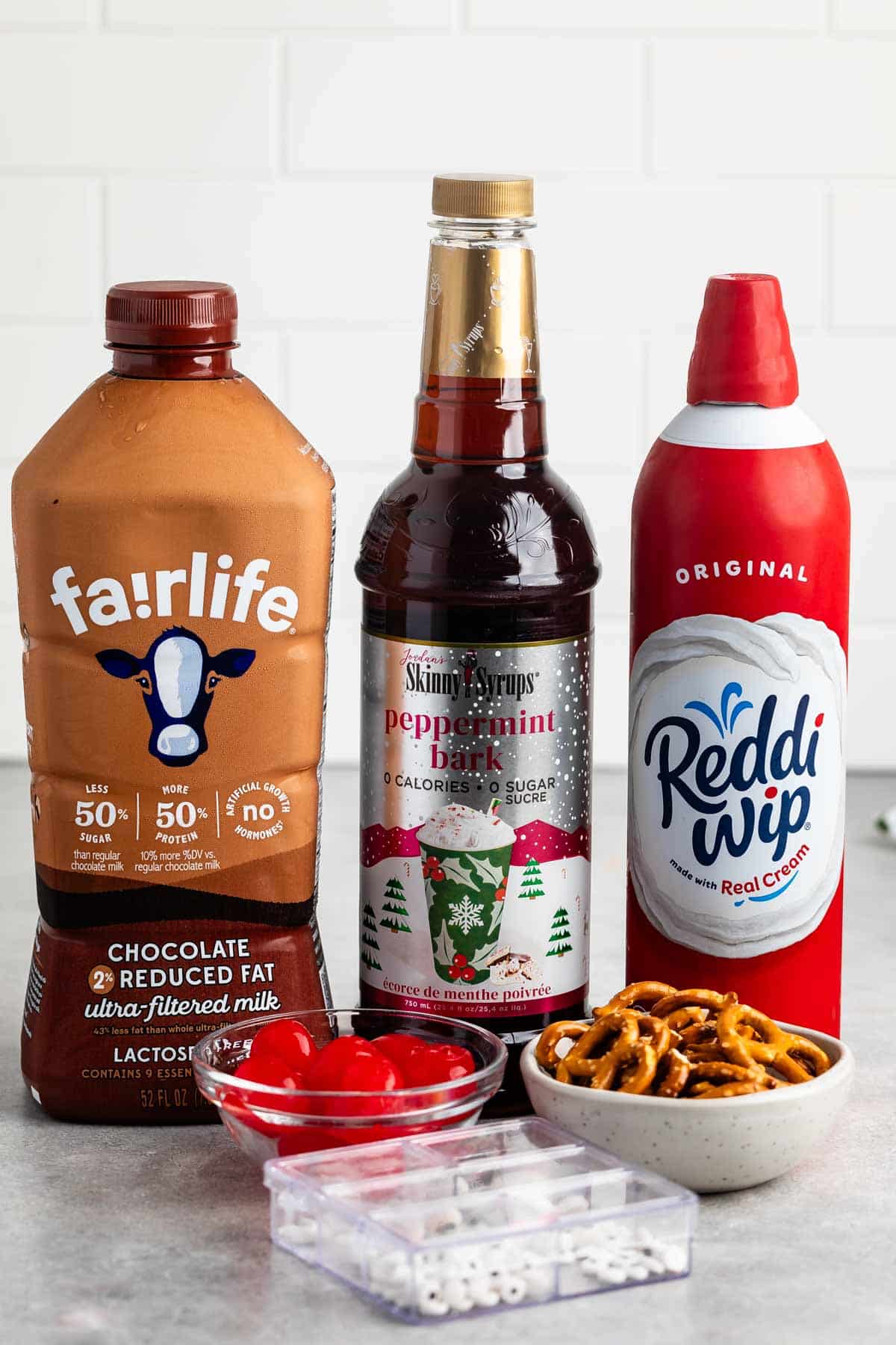 Assorted dessert ingredients on a counter: a bottle of chocolate reduced fat milk, peppermint bark syrup, whipped cream, a bowl of pretzels, a bowl of red cherries, and a small container of white candy eyes.