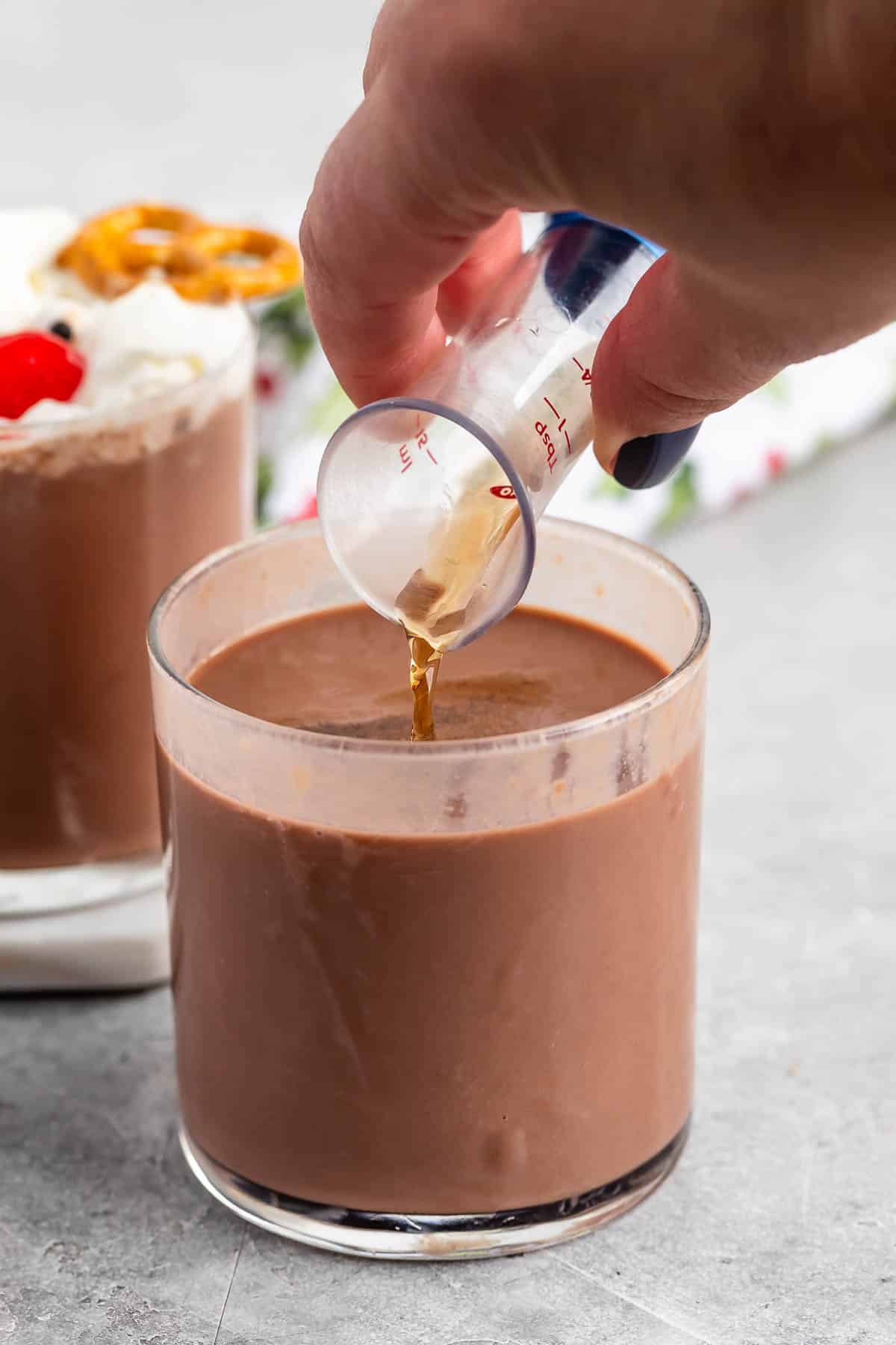 A person pours a small amount of liquid into a glass filled with chocolate milk. Another glass with whipped cream, a cherry, and a pretzel is in the background. The scene is set on a gray surface.