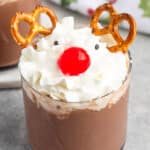 A glass filled with a chocolate drink topped with whipped cream, resembling a reindeer. Pretzels are used for antlers, a red candy for the nose, and two small dots for eyes. A festive cloth is in the background.