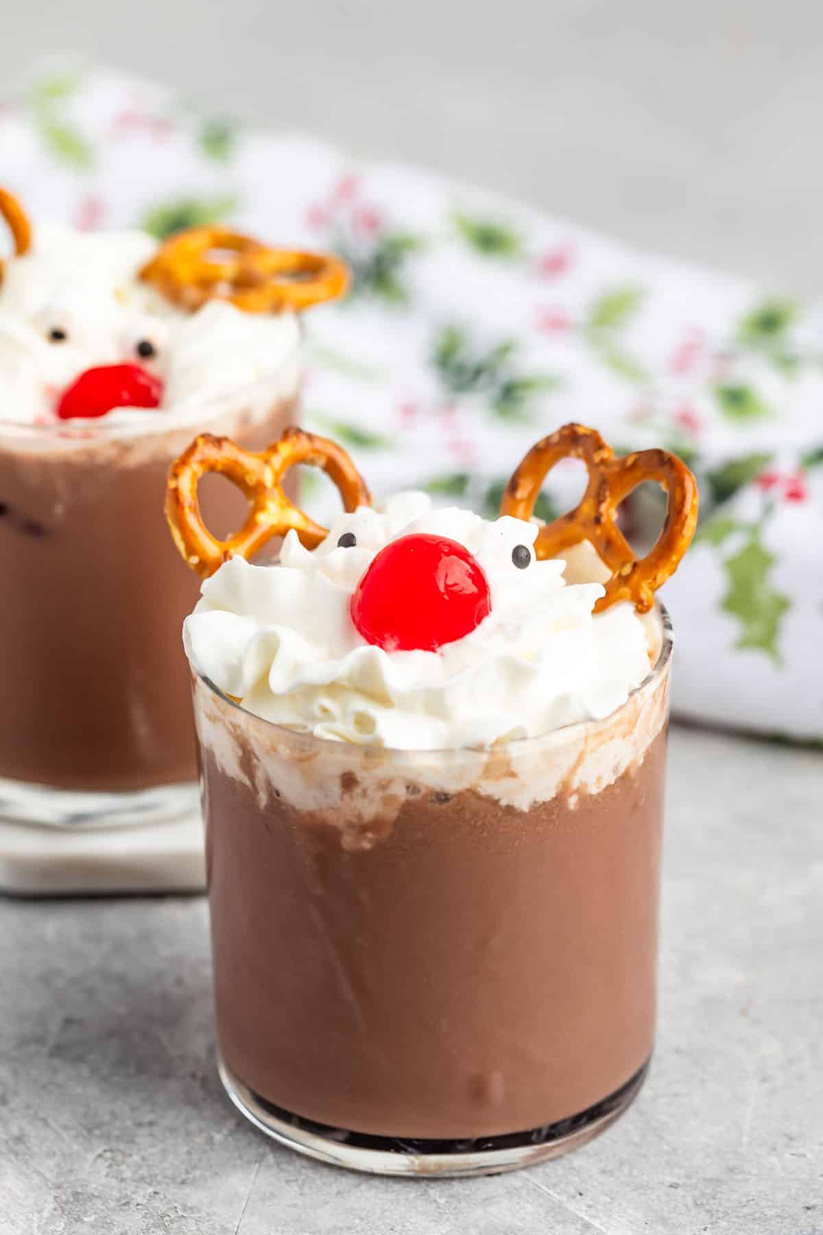 Two glasses of chocolate milk are topped with whipped cream, a cherry, pretzels resembling antlers, and chocolate chips for eyes, resembling reindeer faces. The glasses are set on a light surface with a festive napkin in the background.