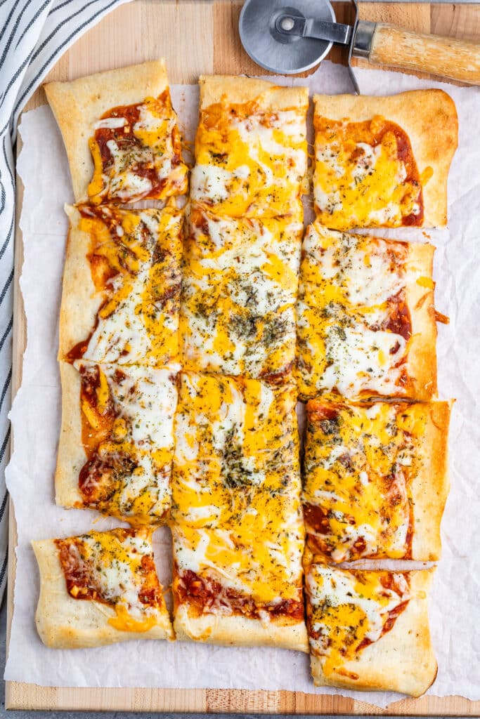 Rectangular pizza with a golden crust topped with melted cheese, herbs, and tomato sauce, cut into square slices. A pizza cutter and striped cloth are visible on a wooden board.