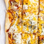 A rectangular pizza topped with melted cheese, herbs, and tomato sauce is sliced into square pieces. The crust is golden brown and crispy, set on parchment paper on a wooden surface.