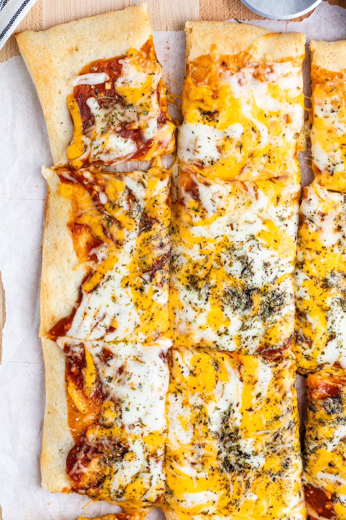 A rectangular pizza topped with melted cheese, herbs, and tomato sauce is sliced into square pieces. The crust is golden brown and crispy, set on parchment paper on a wooden surface.