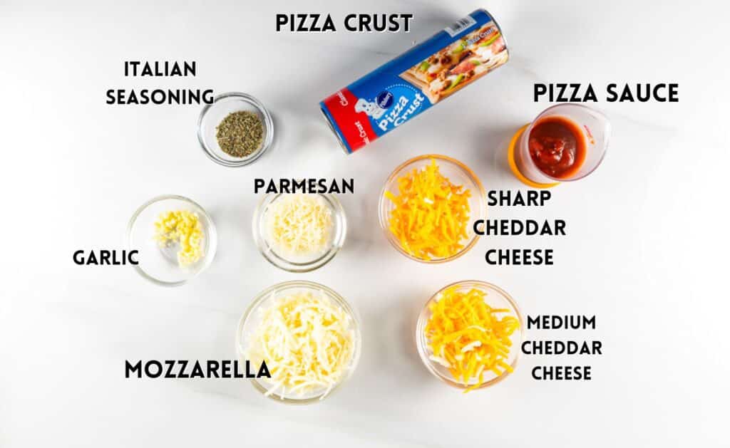 Ingredients for pizza making are arranged on a white surface: a roll of pizza crust, pizza sauce, Italian seasoning, garlic, Parmesan, mozzarella, sharp cheddar cheese, and medium cheddar cheese.