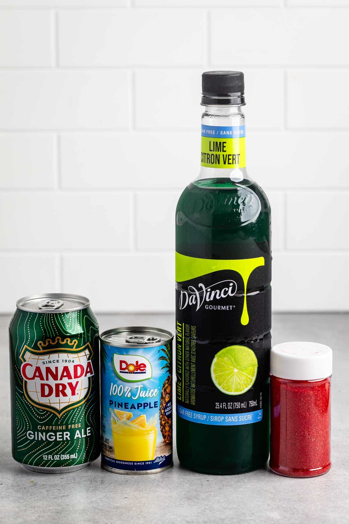 A can of Canada Dry ginger ale, a can of Dole pineapple juice, a bottle of DaVinci Gourmet lime syrup, and a small container of red sugar are arranged on a gray countertop against a white tiled backdrop.