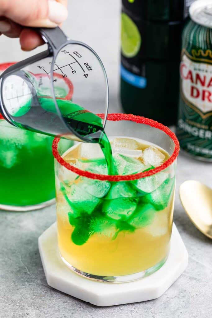 A hand pours green liquid into a glass filled with ice cubes, with a red sugar rim. Another drink and a can of Canada Dry ginger ale are visible in the background, set on a marble surface.