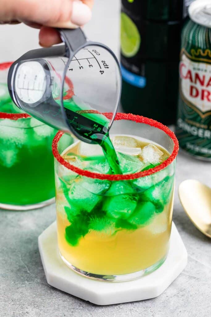 A hand pours green liquid from a measuring cup into a glass filled with ice and a yellow drink. The glass has a red rimming sugar edge. A second green drink, a can of Canada Dry, and a spoon are in the background on a gray surface.