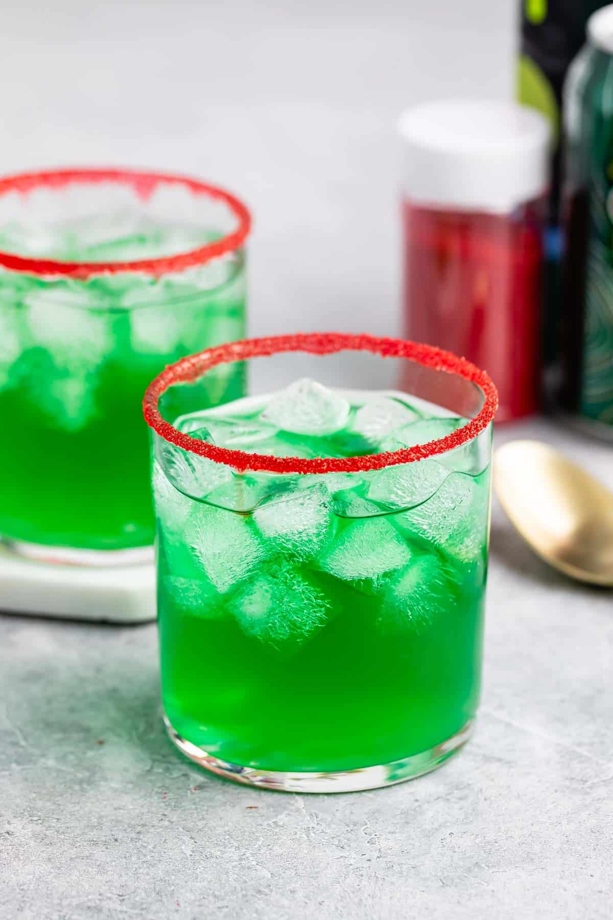 Two glasses of green iced drinks with red sugar rims sit on a light surface. A gold spoon and a container of red sugar are in the background.