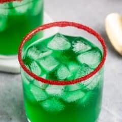 A glass of vibrant green liquid filled with ice cubes, topped with a red sugar rim, is placed on a gray tabletop. Another similar glass and a spoon are blurred in the background.