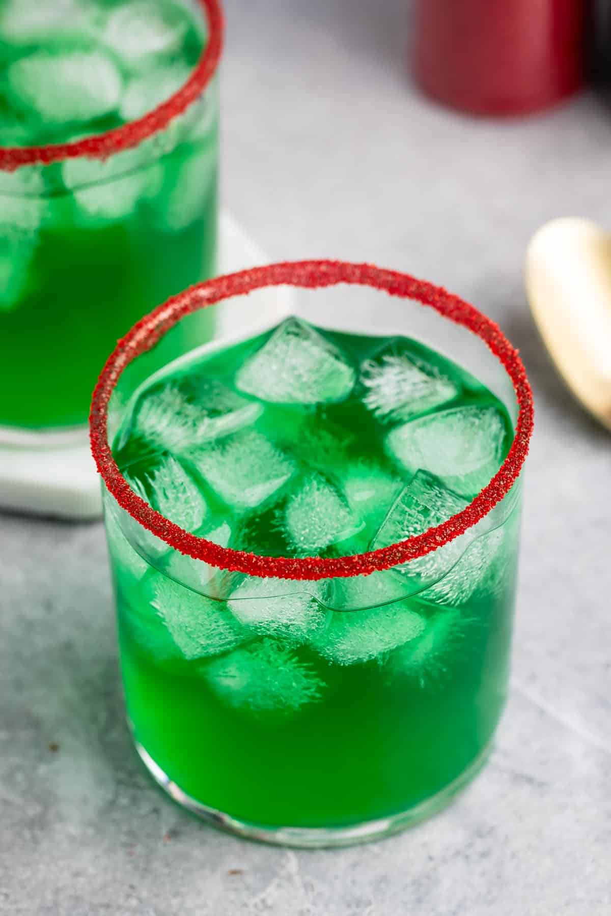 A glass of vibrant green liquid filled with ice cubes, topped with a red sugar rim, is placed on a gray tabletop. Another similar glass and a spoon are blurred in the background.