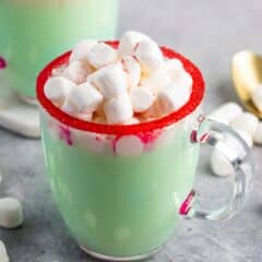 A clear glass mug filled with green hot chocolate is topped with mini marshmallows. The rim of the mug is decorated with red sugar, and a few marshmallows are scattered on the gray surface around it.