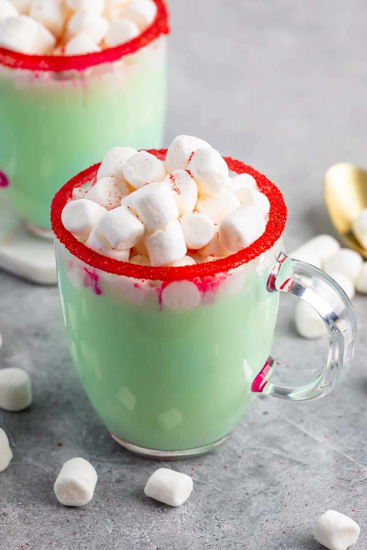 A clear glass mug filled with green hot chocolate is topped with mini marshmallows. The rim of the mug is decorated with red sugar, and a few marshmallows are scattered on the gray surface around it.