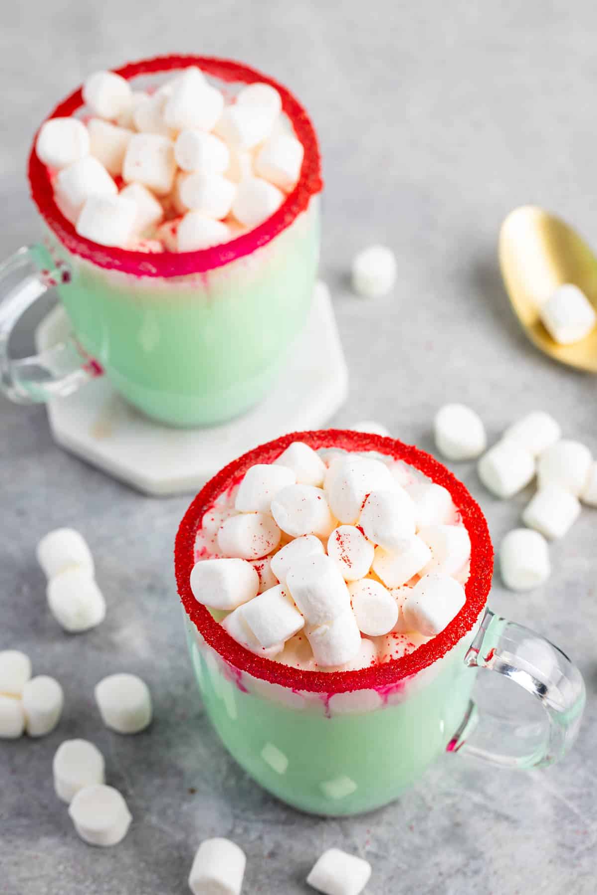 Two glass mugs filled with mint green hot chocolate topped with mini marshmallows. The rims are coated with red sugar. A few marshmallows are scattered around on a gray surface, with a gold spoon nearby.