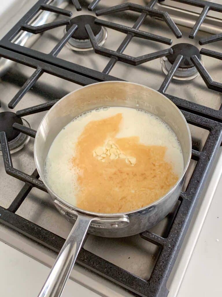 A saucepan on a stove contains simmering milk with melted butter and chocolate chips. The stove has a metal grate with four burners.