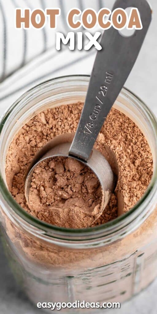 A jar of hot cocoa mix with a metal measuring scoop resting in the powder. The top of the image displays the text Hot Cocoa Mix in bold letters. The bottom shows the website easygoodideas.com.