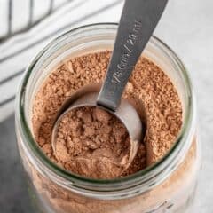 A glass jar filled with cocoa powder is on a countertop. A metal 1/8 cup measuring scoop is resting inside the jar. A striped cloth is partially visible in the background.