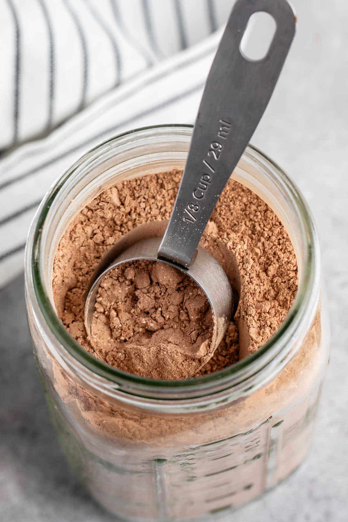 A glass jar filled with cocoa powder is on a countertop. A metal 1/8 cup measuring scoop is resting inside the jar. A striped cloth is partially visible in the background.