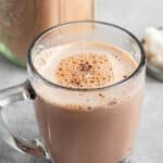 A clear glass mug filled with creamy hot chocolate, topped with a light froth and specks of cocoa. It sits on a gray surface near a jar and some marshmallows.