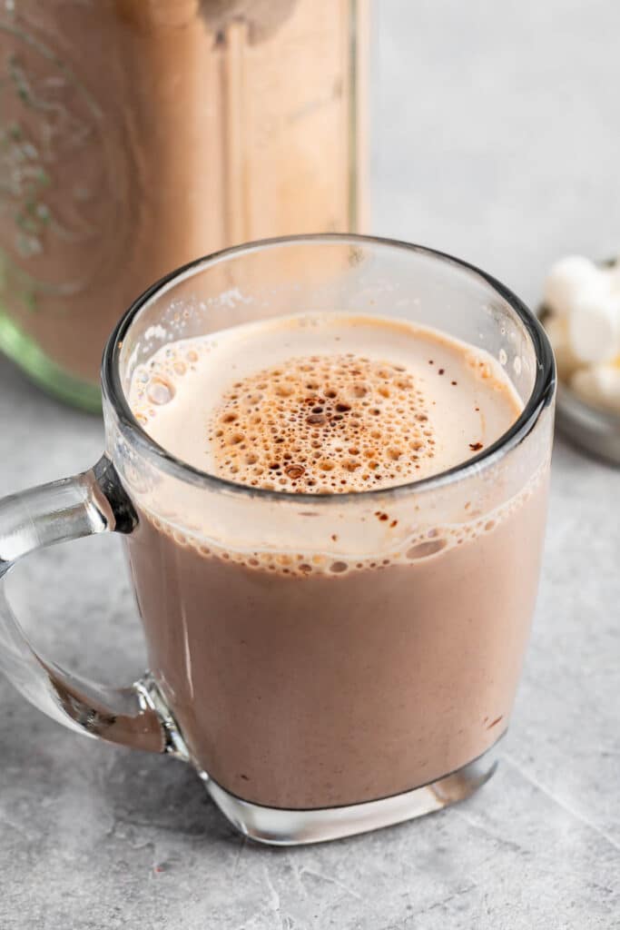 A clear glass mug filled with creamy hot chocolate, topped with a light froth and specks of cocoa. It sits on a gray surface near a jar and some marshmallows.