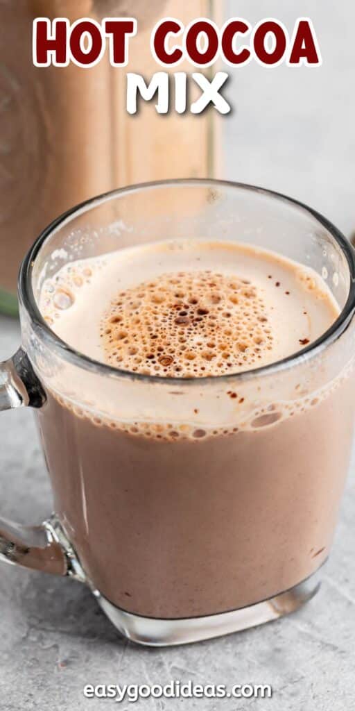 A clear glass mug filled with frothy hot cocoa sits on a gray surface. The words HOT COCOA MIX are written at the top in bold red and white letters. A website name is visible at the bottom of the image.