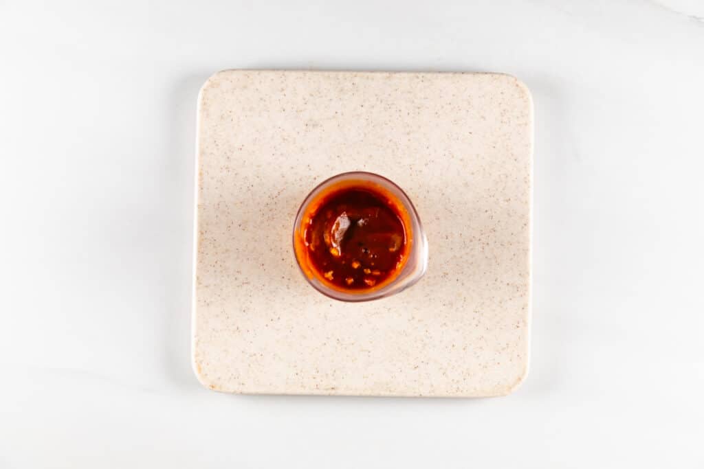 A small glass filled with red chili oil, including visible chili flakes, is placed on a speckled square plate against a white background.